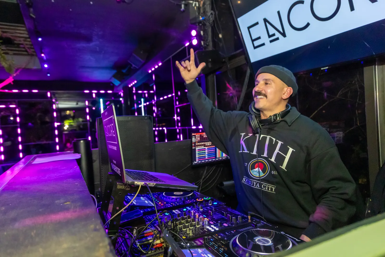 A DJ throwing a hand up in the booth under purple neon